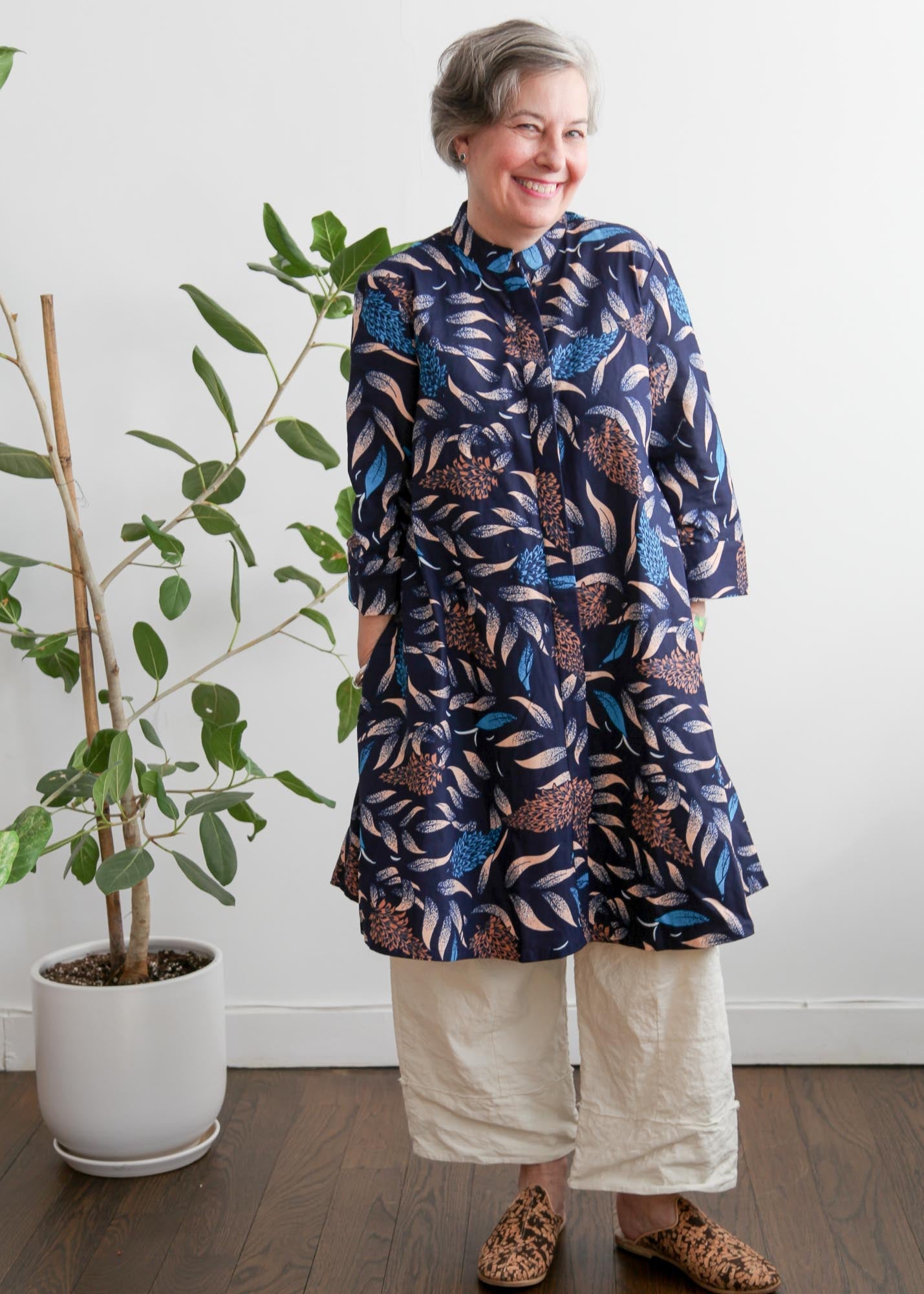 A woman standing next to a potted plant, wearing a navy, aqua, and apricot floral &amp; leaf print dress with button fastening through the front.