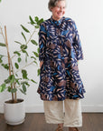 A woman standing next to a potted plant, wearing a navy, aqua, and apricot floral & leaf print dress with button fastening through the front.